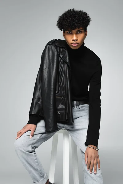 african american man with leather jacket on shoulder looking at camera while sitting isolated on grey