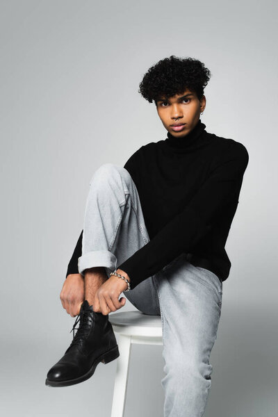 trendy african american man in black turtleneck and jeans sitting on white stool and looking at camera on grey background