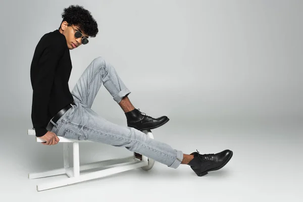 身穿黑色高领毛衣 牛仔裤和深色太阳镜的时髦的非洲裔美国男人坐在翻倒的灰色凳子上 — 图库照片
