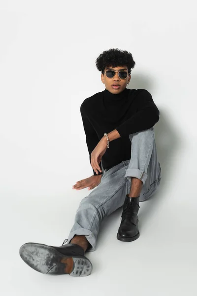 young african american man in jeans and black turtleneck looking at camera while sitting on white