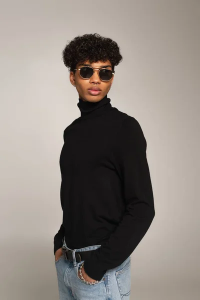 african american man in black turtleneck and sunglasses standing with hands in pockets of jeans isolated on grey