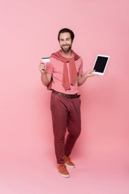 Elinde tablet, elinde boş ekran ve pembe arka planda kredi kartıyla gülümseyen adamın tam uzunluğu 