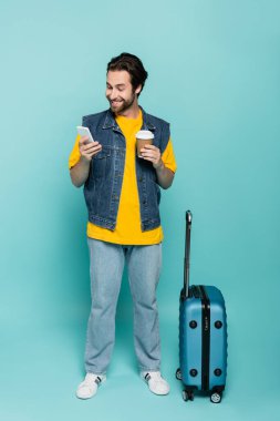 Kahveli gülümseyen adam mavi arka planda bavulun yanında akıllı telefon kullanıyor. 