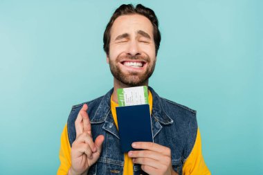 Gözleri kapalı gülümseyen bir adam pasaport ve uçak biletini elinde tutarken mavi üzerinde parmaklarını çapraz yapıyor. 