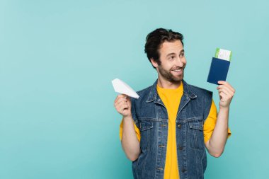 Elinde kağıt uçak ve pasaportu olan gülümseyen bir adam mavi üzerine izole edilmiş uçak bileti taşıyor. 