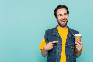 Young man sticking out tongue while pointing at coffee to go isolated on blue clipart