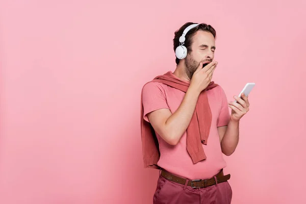 ピンクの上に隔離されたスマートフォンを保持しながら ヘッドフォンYawningで若い男 — ストック写真