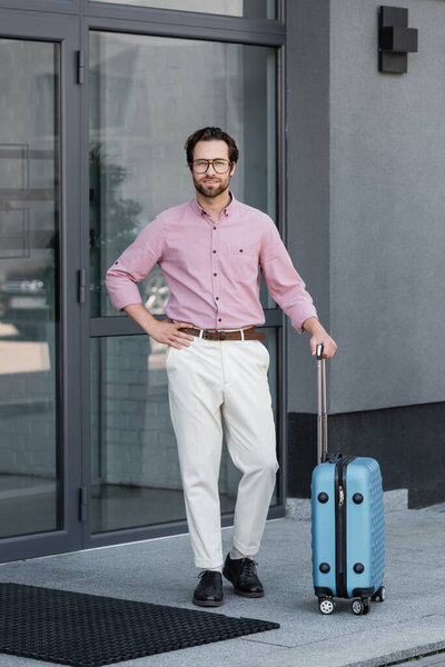Smiling businessman with suitcase standing near building 