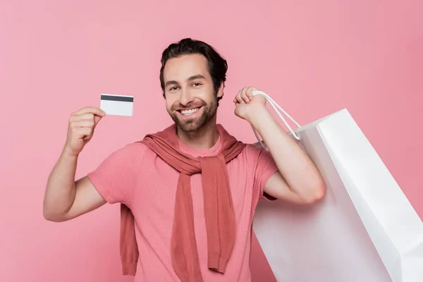 持用粉红隔离的购物袋时出示信用卡的年轻人 — 图库照片
