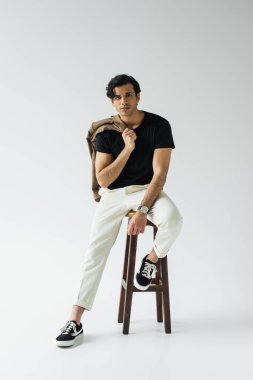 Gri sandalyede otururken spor ceket tutan genç bir adam.