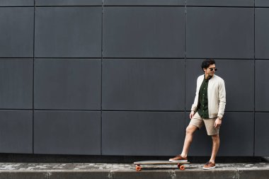 Binanın yanında güneş gözlüklü, uzun sörf tahtası olan uzun boylu şık bir adam. 