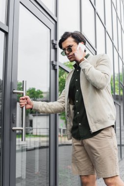 Güneş gözlüklü şık genç adam binaya girerken cep telefonuyla konuşuyor. 