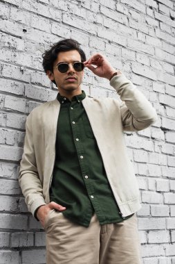 young man in stylish sunglasses and bomber jacket posing with hand in pocket near brick wall clipart