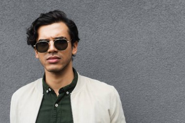 young man in stylish sunglasses and bomber jacket posing near grey wall clipart