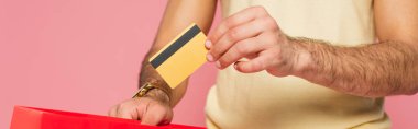 cropped view of young man putting credit card in red shopping bag isolated on pink, banner clipart