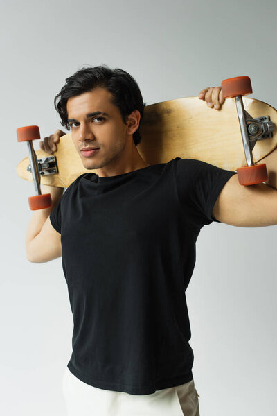 young man in black t-shirt holding longboard and looking at camera isolated on grey