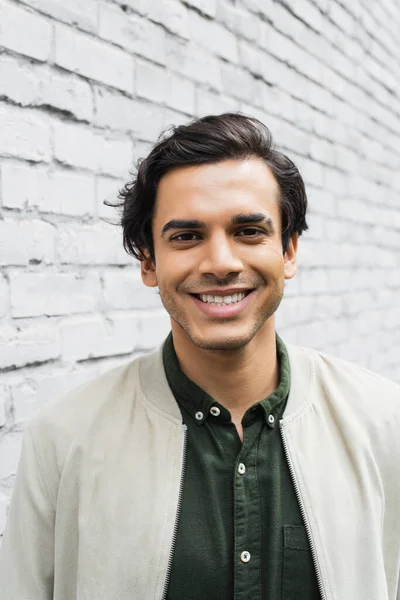 happy young man in bomber jacket smiling near brick wall