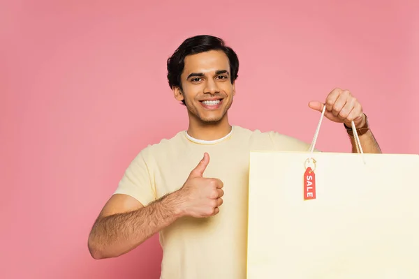 笑顔の若い男がショッピングバッグを持って販売タグと親指を示すピンクに隔離された — ストック写真