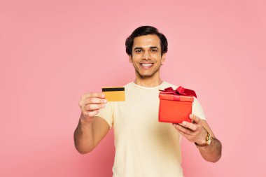 Genç, neşeli bir adam elinde kırmızı bir hediye kutusu ve pembe bir kredi kartı tutuyor.