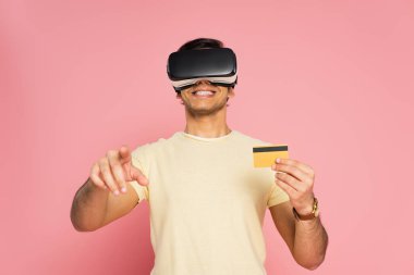 VR kulaklıklı neşeli bir adam kredi kartı tutuyor ve parmağıyla pembe olanı işaret ediyor.