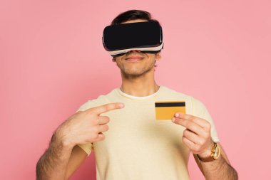 VR kulaklıklı neşeli adam, pembe kartla izole edilmiş kredi kartını işaret ediyor.
