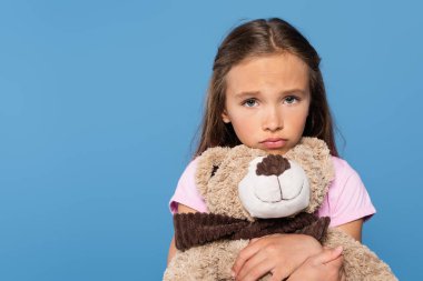 Sad girl holding soft toy and looking at camera isolated on blue  clipart