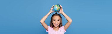 Smiling child holding globe above head isolated on blue, banner  clipart
