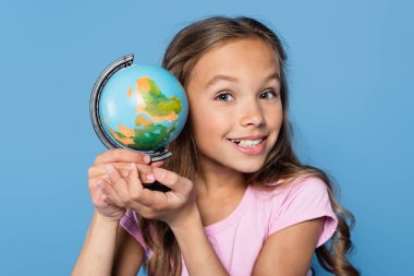 Okul çocuğu dünyayı tutuyor ve mavi üzerine izole edilmiş kameraya bakıyor 