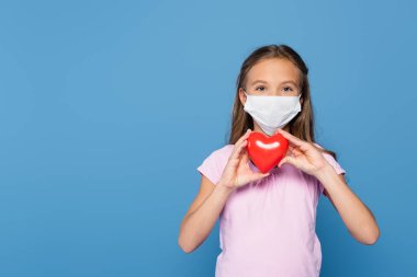 Child in medical mask holding red heart isolated on blue  clipart