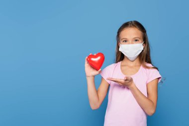 Child in medical mask pointing at decorative heart with hand isolated on blue  clipart