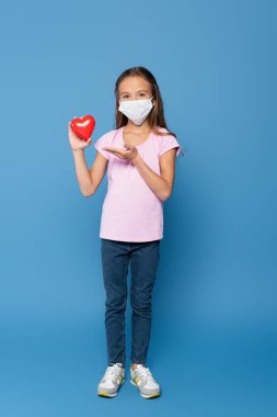 Girl in protective mask holding red decorative heart and pointing with hand on blue background  clipart