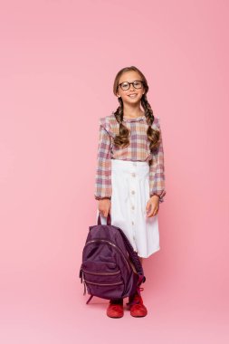Pembe arka planda sırt çantasıyla kameraya gülümseyen şık bir öğrenci. 