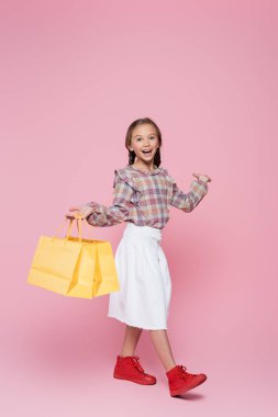 Elinde alışveriş torbaları olan gülümseyen çocuk pembe arka planı işaret ediyor. 