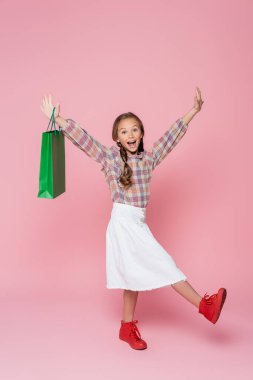 excited girl with green shopping bag raising hands on pink background clipart