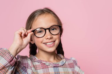 joyful kid in checkered blouse adjusting eyeglasses isolated on pink clipart