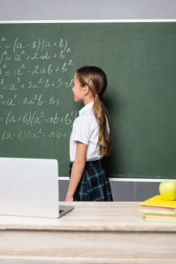 Liseli kız denklemlerle tahtaya yakın duruyor ve dizüstü bilgisayarı olan bir masa. 
