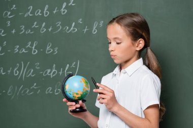 thoughtful schoolkid holding pen while looking at globe near chalkboard clipart