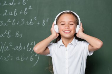 Gözü kapalı neşeli kız kulaklıkla kara tahtanın yanında müzik dinliyor.