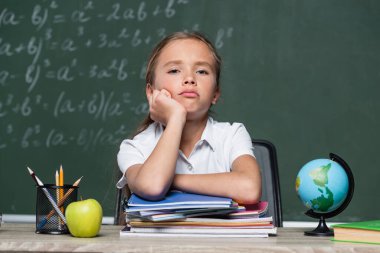 üzgün liseli kız, arka plandaki not defterlerinin, dünya ve karatahtanın yanındaki kameraya bakıyor.