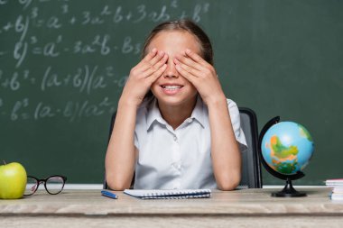 cheerful schoolgirl covering eyes with hands near globe and notebook on desk clipart