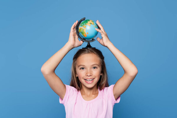 Kid smiling at camera while holding globe on head isolated on blue