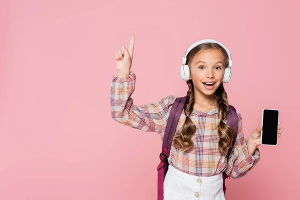 ピンクに隔離されたスマートフォンを保持しながら アイデアを持つヘッドフォンの正の学童 — ストック写真