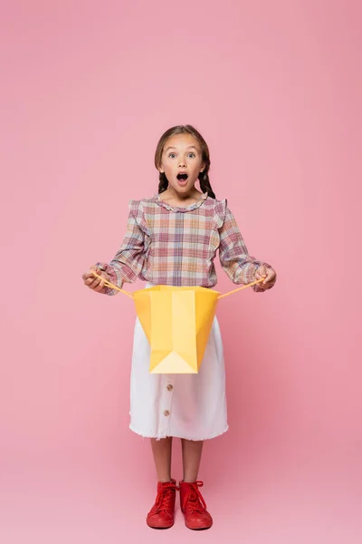 ピンクの背景に黄色のショッピングバッグを開きながらカメラを見て驚く子供 — ストック写真