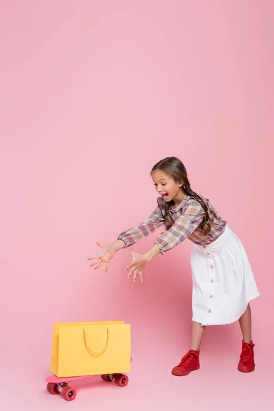 興奮した女の子とともにオープン口と手近くに黄色ショッピングバッグとペニーボード上のピンクの背景 — ストック写真