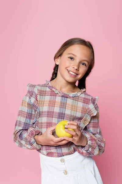 穿着格子花衬衫的快乐女孩拿着新鲜的苹果 看着被粉色隔离的相机 — 图库照片