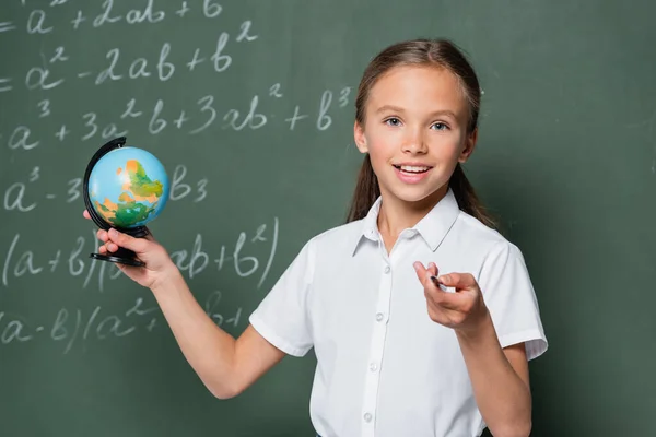 小さな球体を持った幸せな小学生は方程式の付いた黒板の近くのカメラで鉛筆で指します — ストック写真