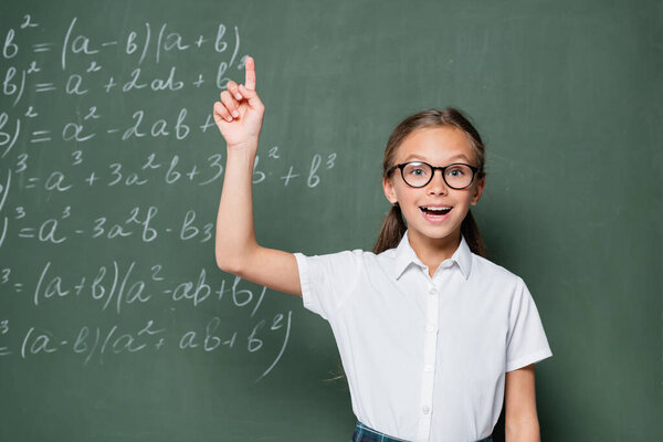 excited schoolgirl in eyeglasses showing idea gesture near chalkboard with equations