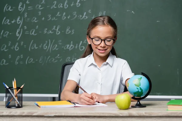 Leende Skolflicka Glasögon Skriva Anteckningsbok Nära Jordklotet Och Suddig Tavlan — Stockfoto