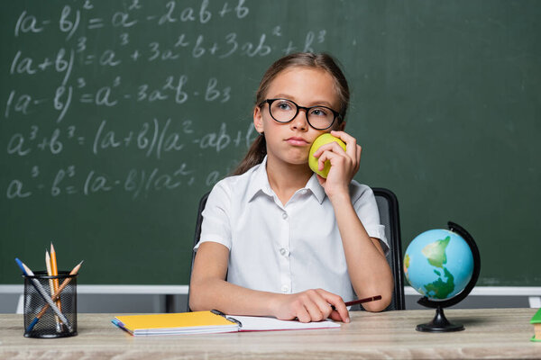 displeased schoolgirl holding apple near globe and notebook on desk