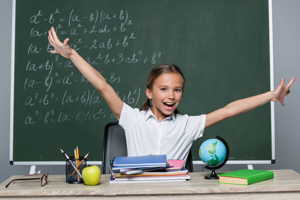 cheerful schoolgirl with open arms near notebooks on desk and chalkboard with equations